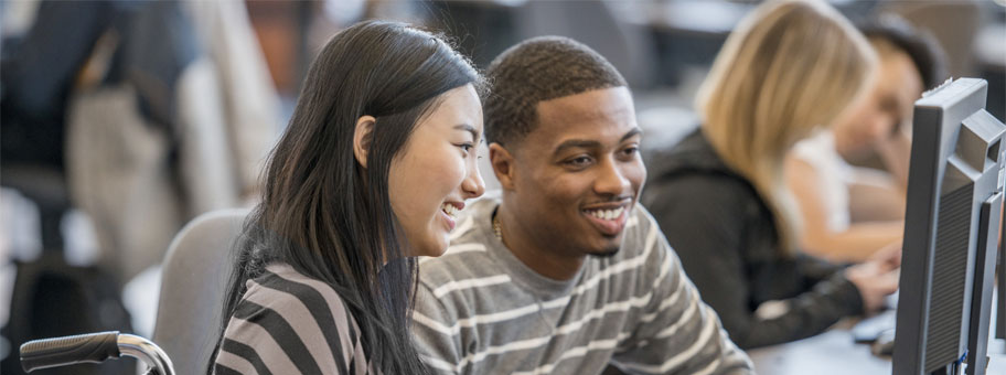 Two students sit in front of a computer monitor smiling.